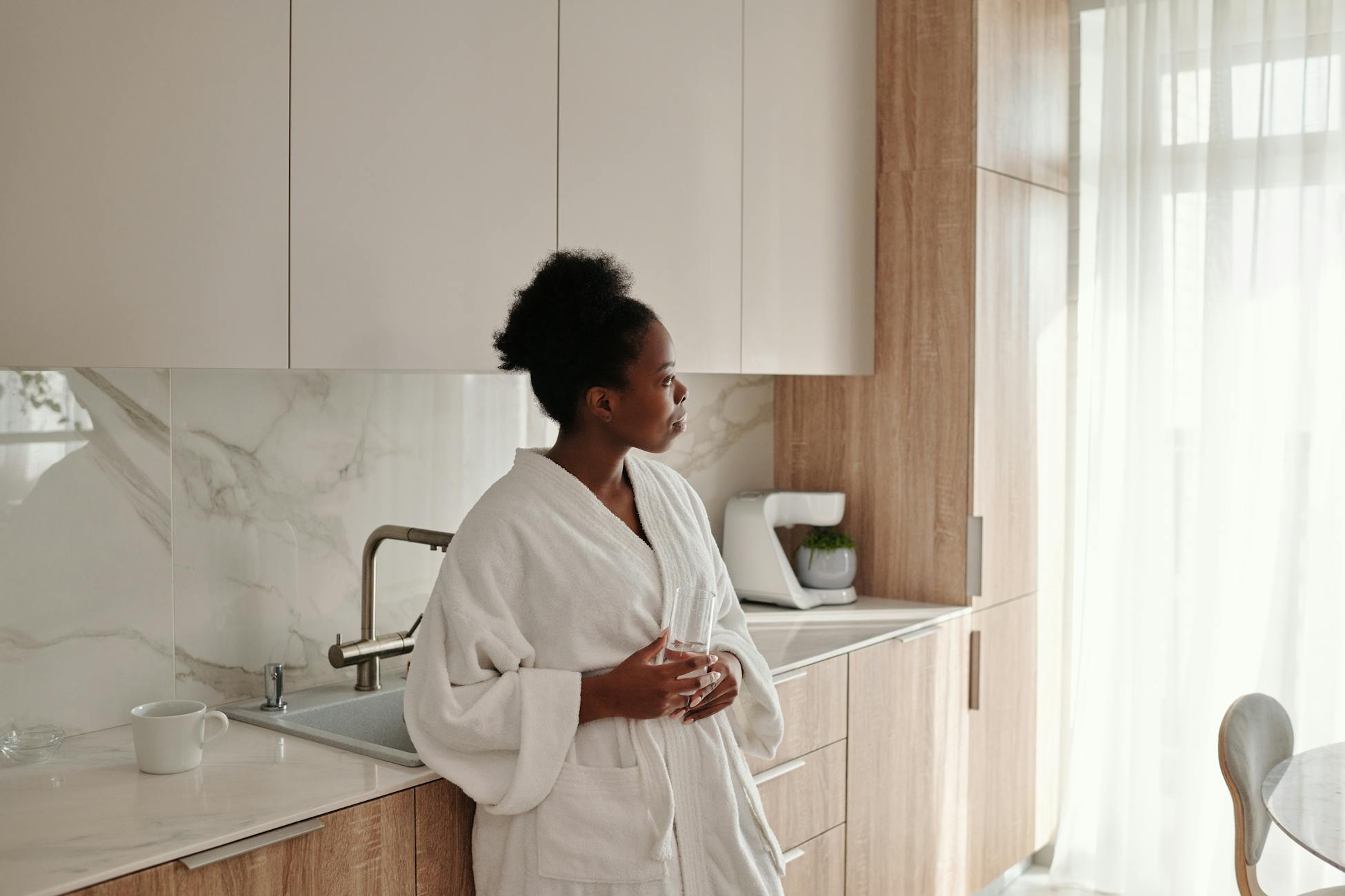 Black woman in bathrobe drinking coffee and looking out window in bright kitchen.