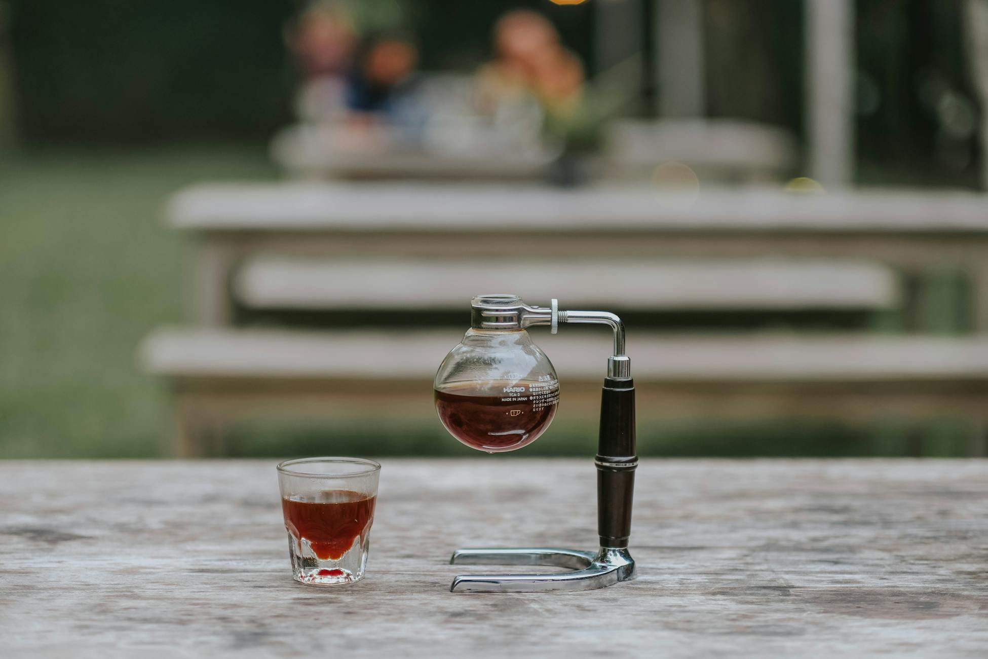 Close-up of a siphon coffee maker and coffee cup on a wooden table outdoors.
