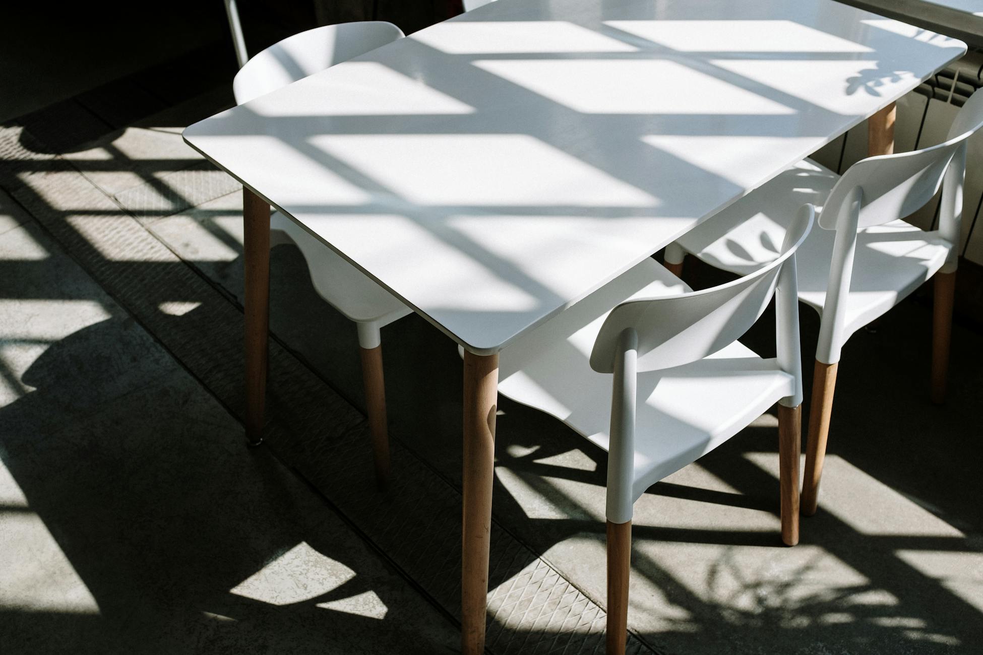 A minimalist setup of white table and chairs with sunlight streaming through a window casting shadows indoors.
