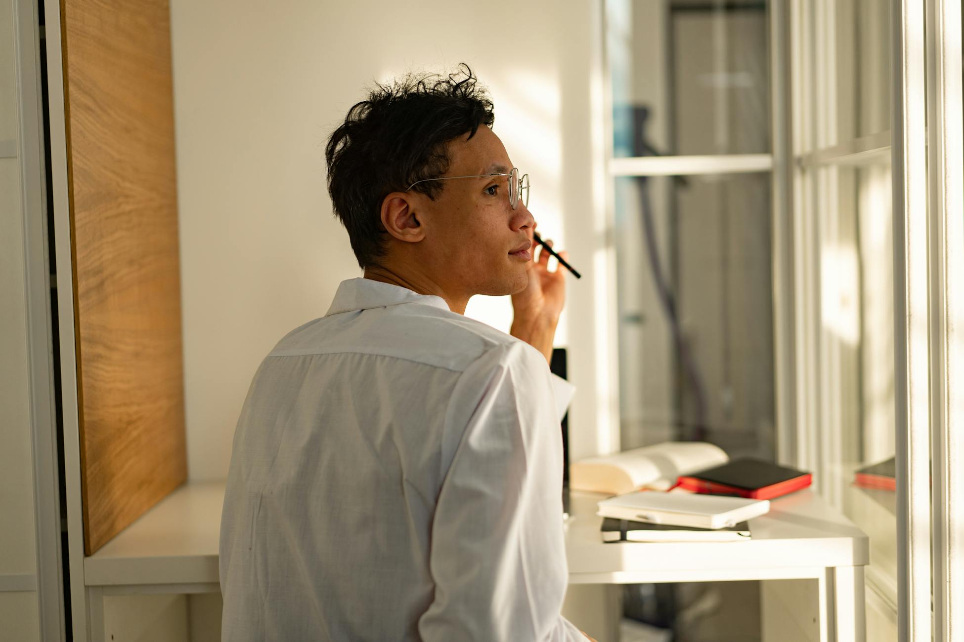 Man in white shirt gazing out window pensively, surrounded by books in modern office.