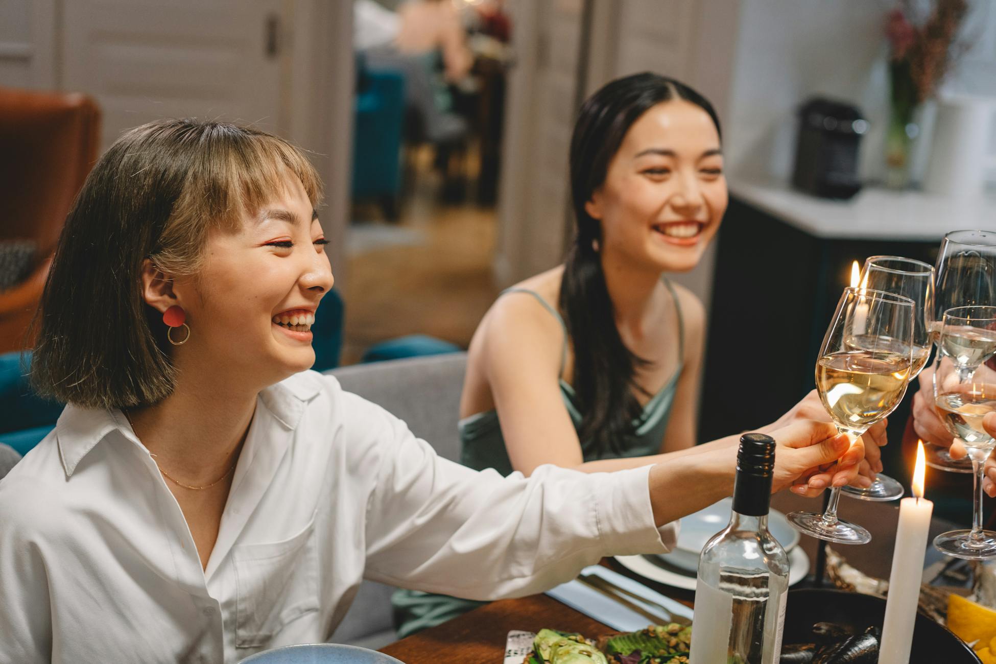 Two women enjoying a lively dinner party, clinking glasses of white wine in a cozy setting.