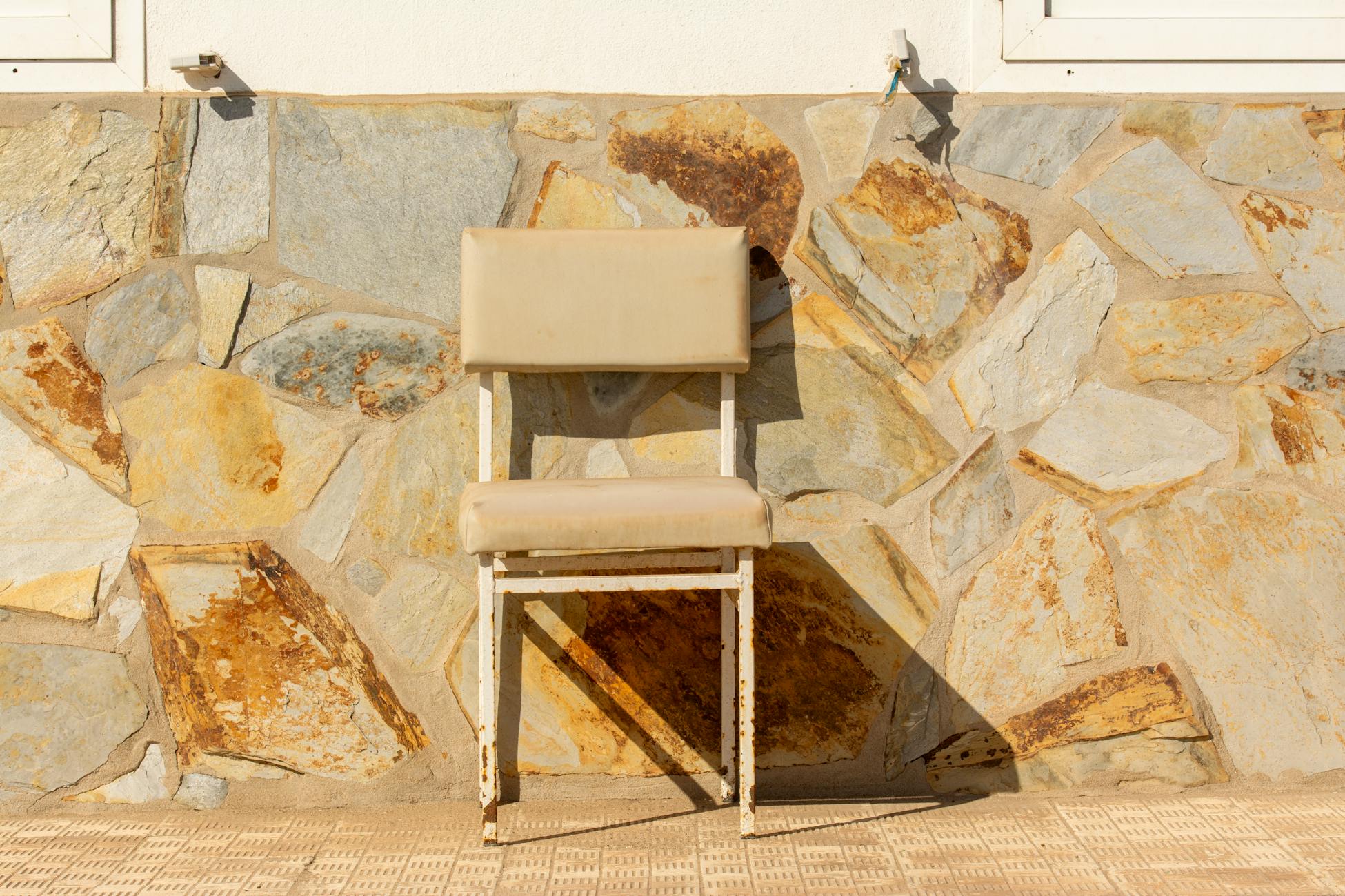 A solitary rustic chair in bright sunlight against a textured stone wall, evoking urban simplicity and warmth.
