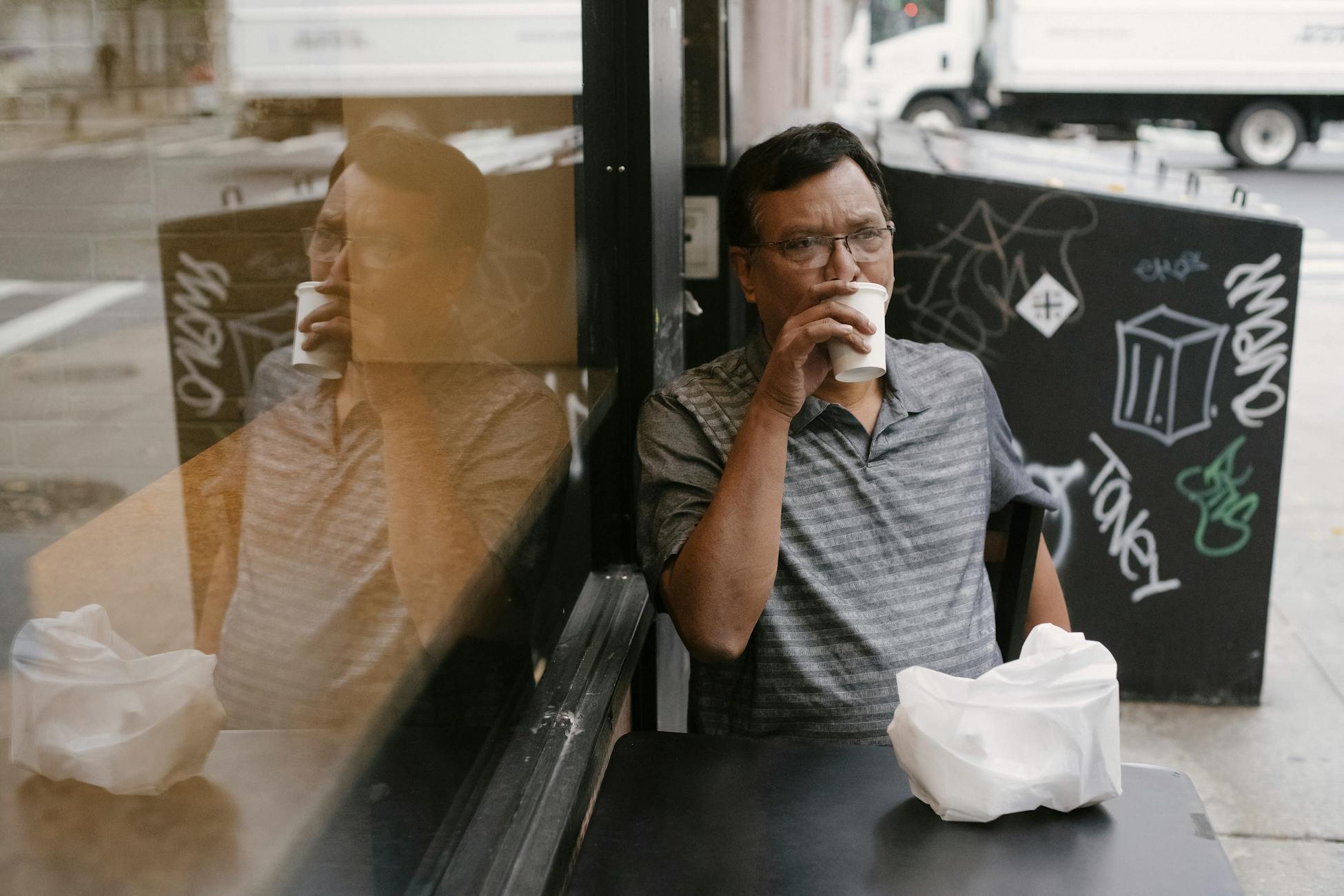 Pensive Asian male in eyeglasses sitting at table in street cafeteria while drinking hot beverage from paper cup on street in city