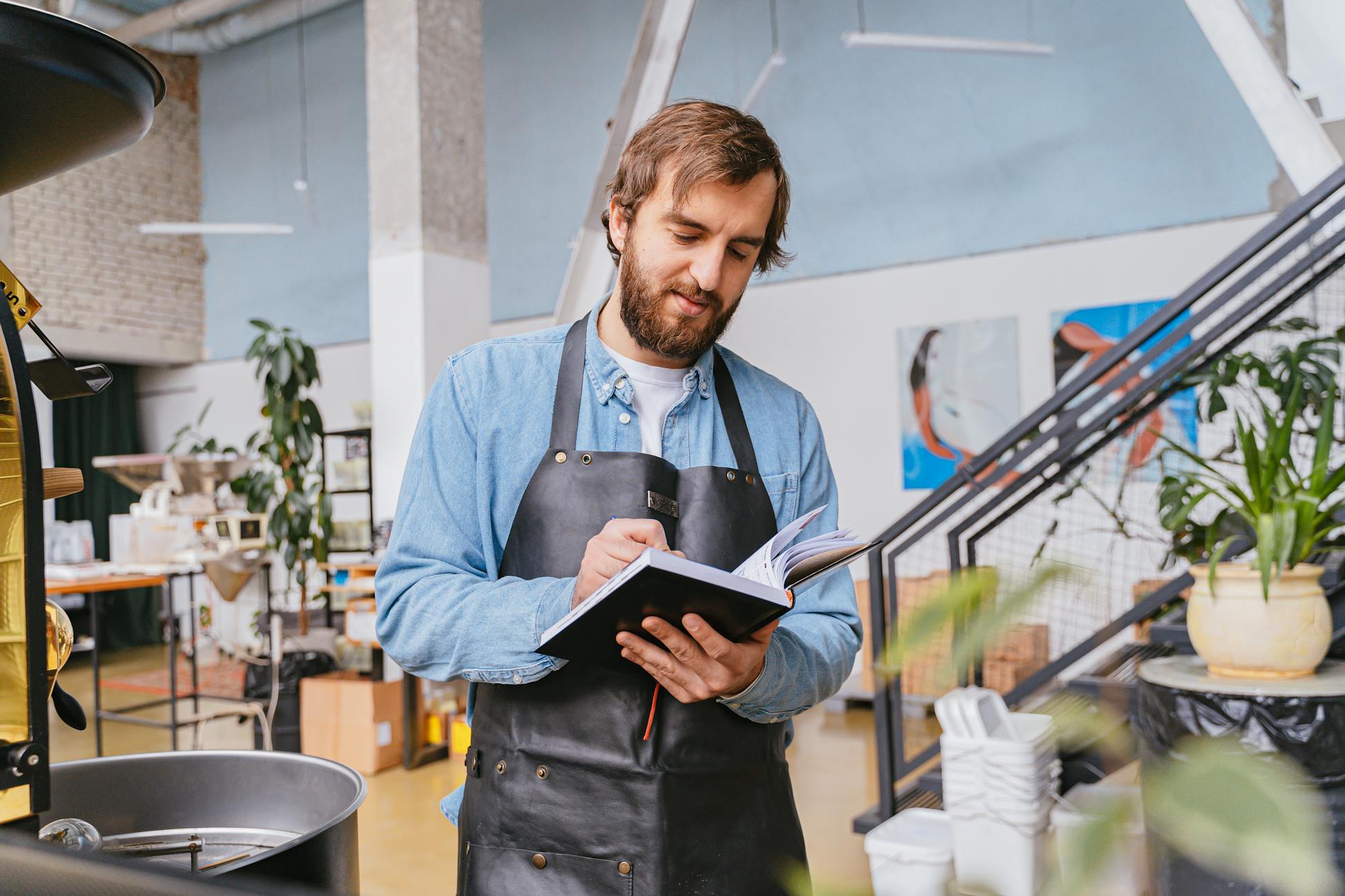 Bearded man in an apron writing in a notebook in a modern cafe setting.