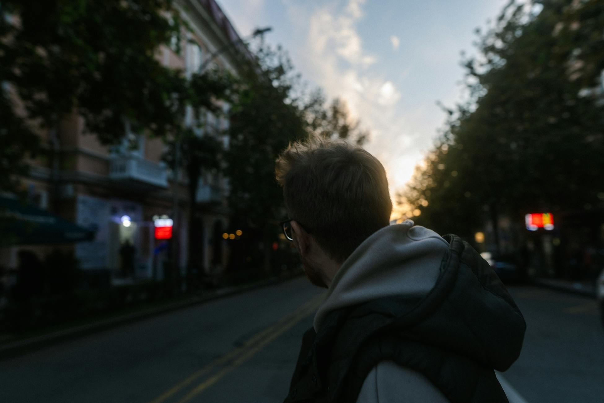 A man in a hoodie walks down a tree-lined city street during sunset.