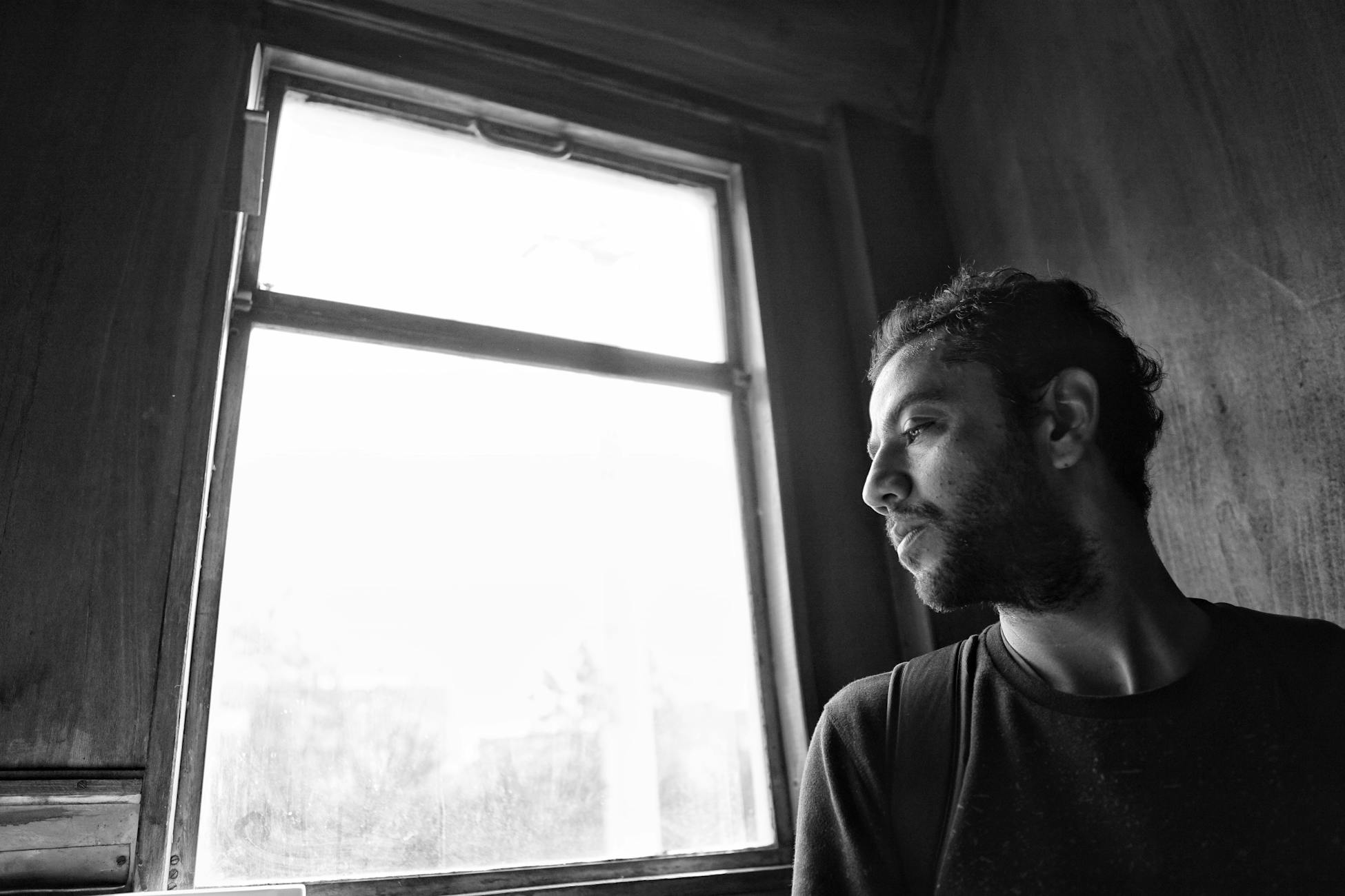 A contemplative man looks out of a window in a black and white photograph, evoking thoughtfulness.