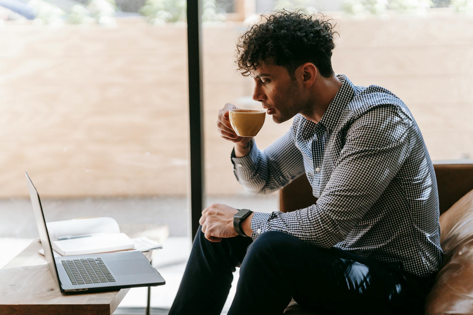 A man in smart casual attire drinks coffee while using a laptop in a modern indoor setting.