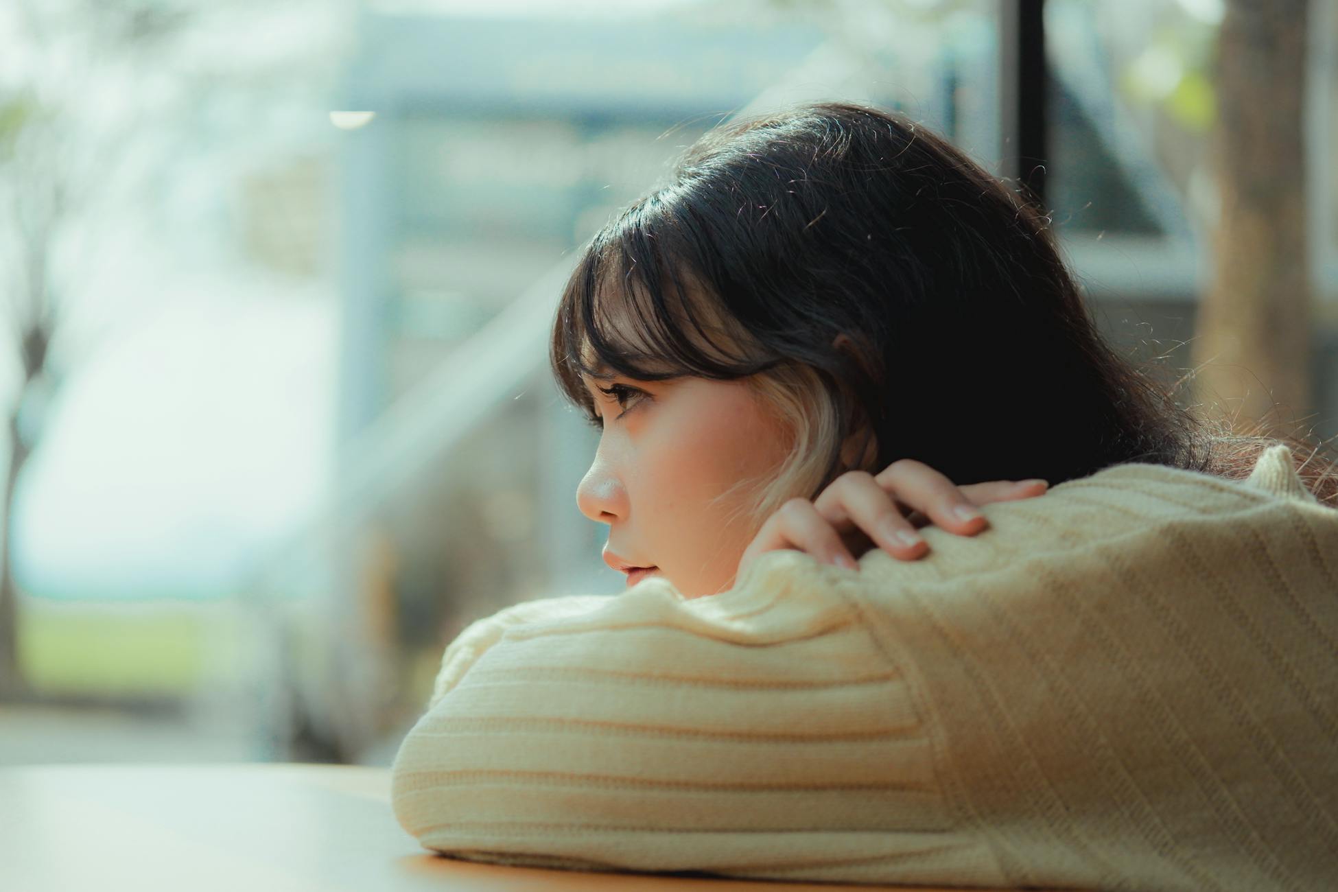 A woman leans thoughtfully on a table by a window, enjoying a moment of relaxation indoors.