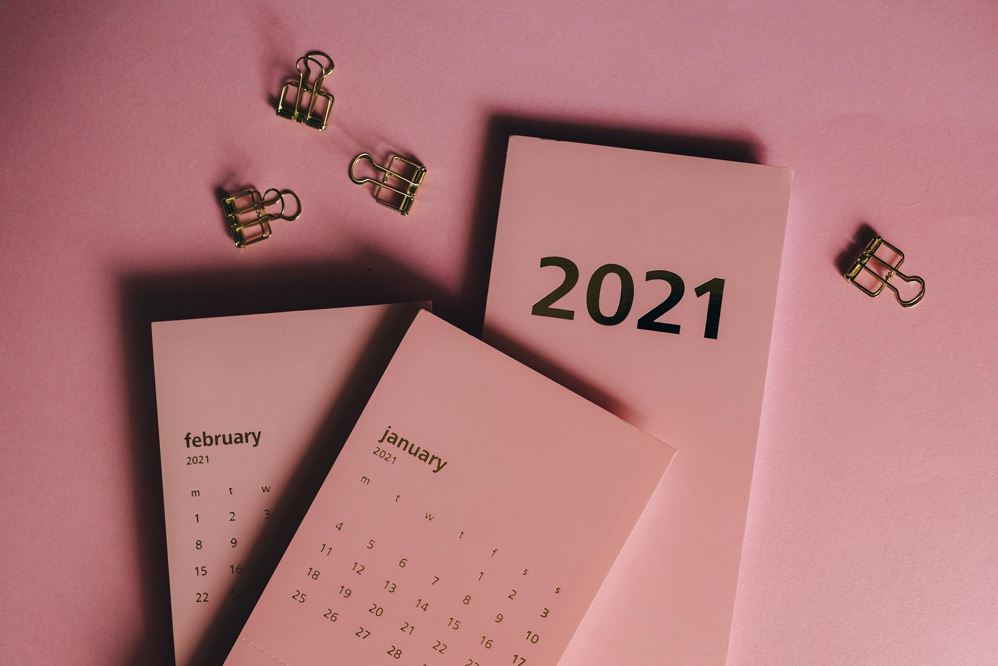 Top view of modern paper calendars with numbers and words near metal clips on pink background