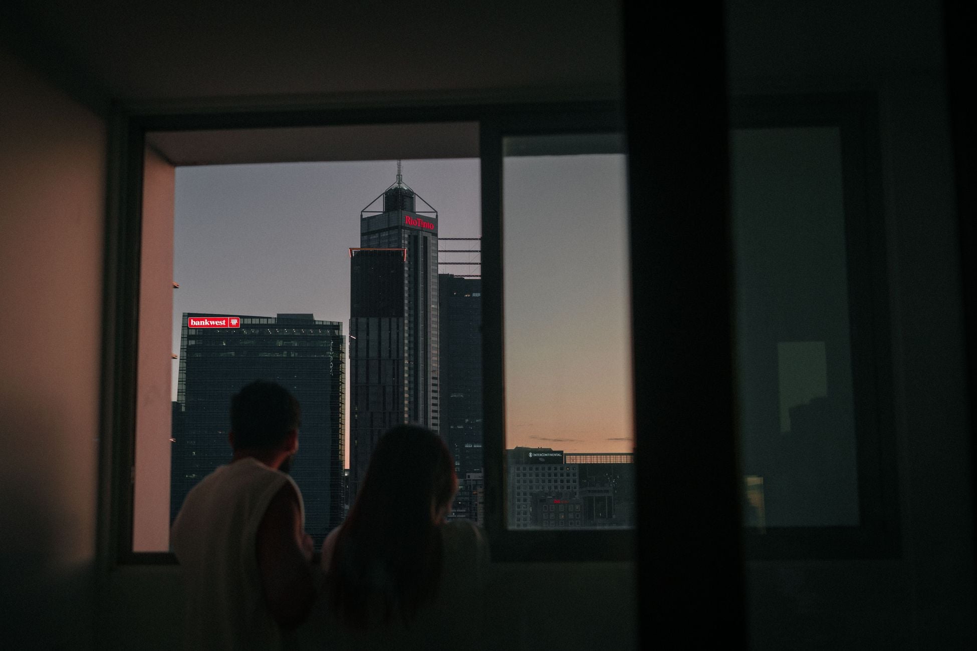 Silhouette of a couple looking out at skyscrapers through a window during sunset.