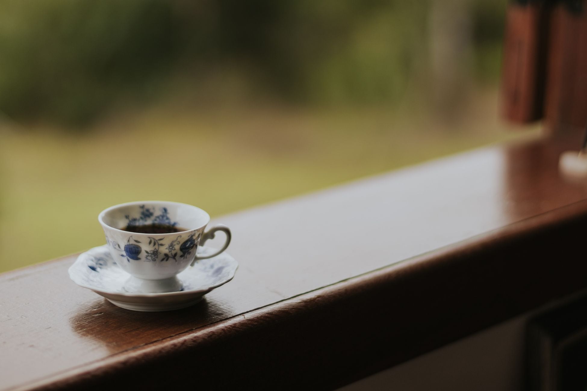 A delicate porcelain cup and saucer with floral design on a wooden windowsill, with a blurred green background.