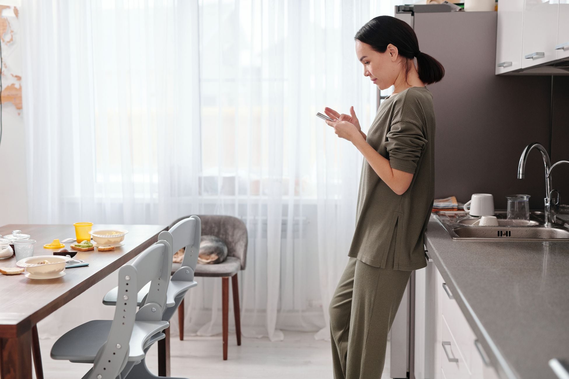 Woman standing in kitchen using phone, morning light, casual home scene.
