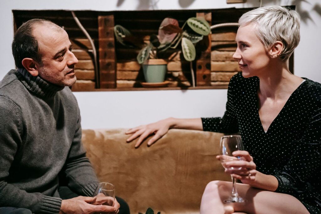 Side view of couple sitting on sofa with glasses of drink and having conversation