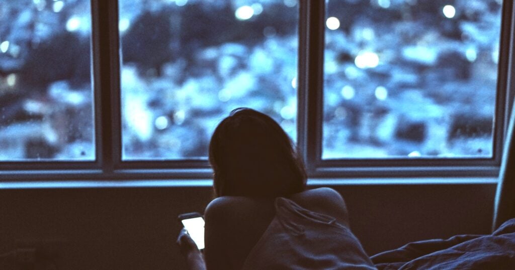 A person sits alone at a window at dusk, seen from behind, holding a lit phone screen in one hand while city lights blur outside.